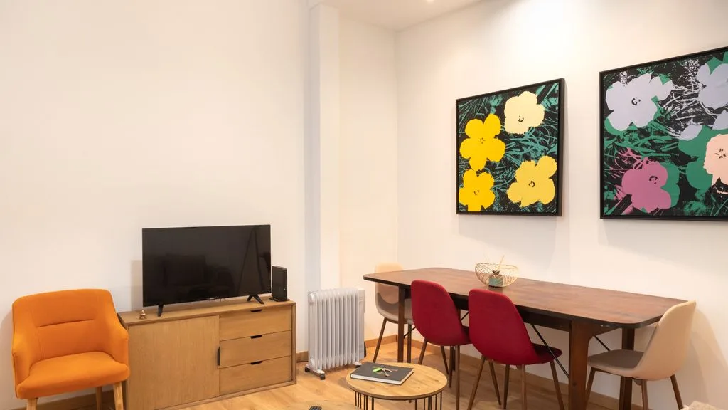 Comedor con mesa de madera, sillas de colores y cuadros florales en la pared.