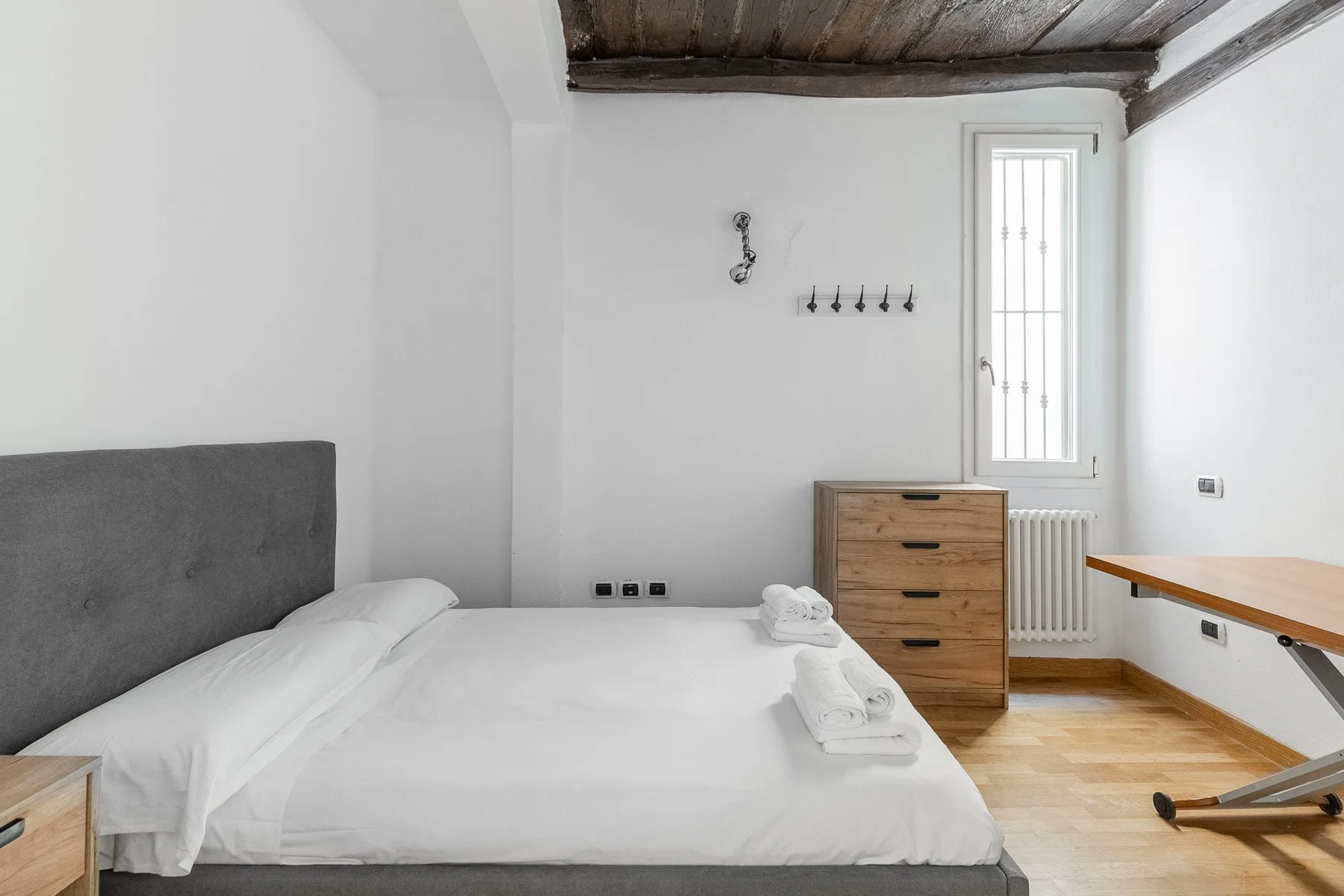 A bright bedroom with a white bed, wooden dresser, and narrow window.