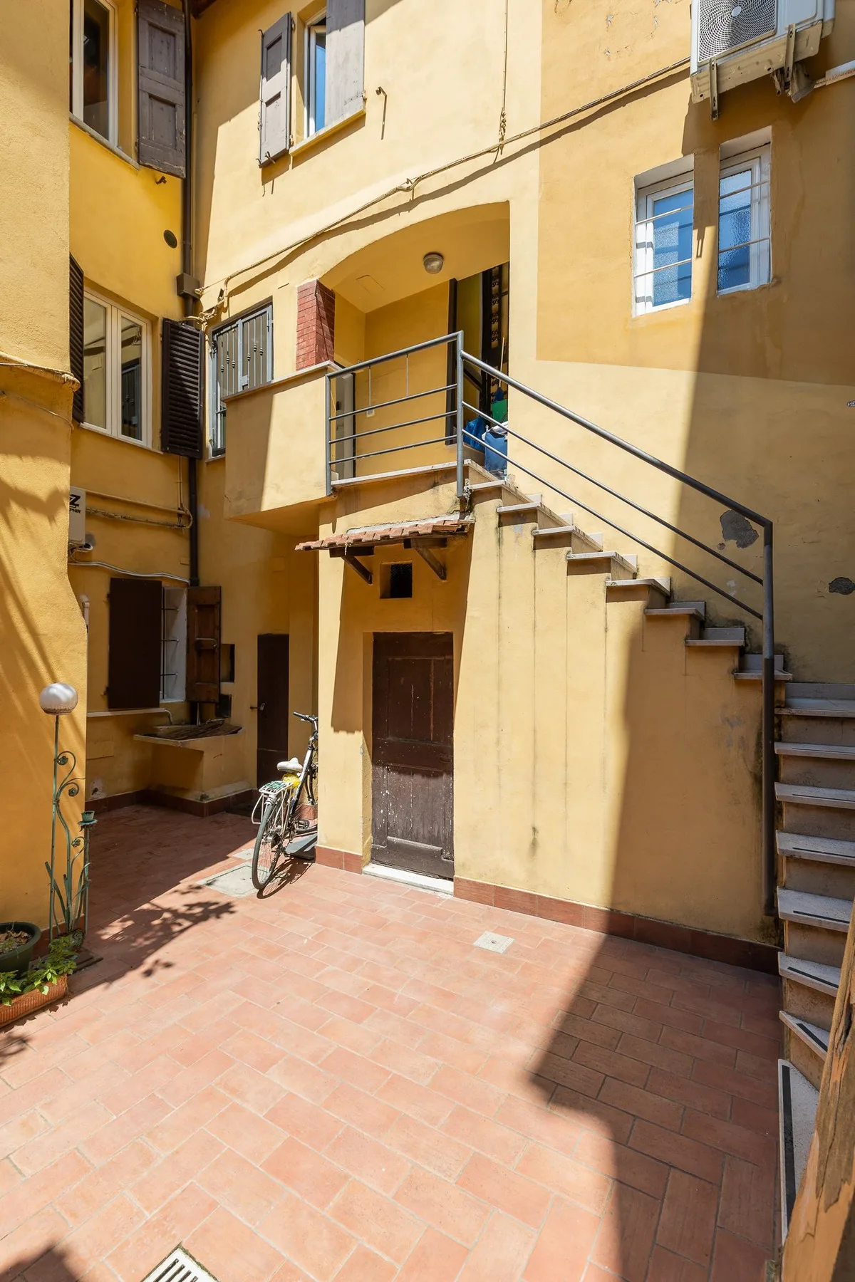 A sunlit courtyard with an exterior staircase and a bicycle beside the doorway.