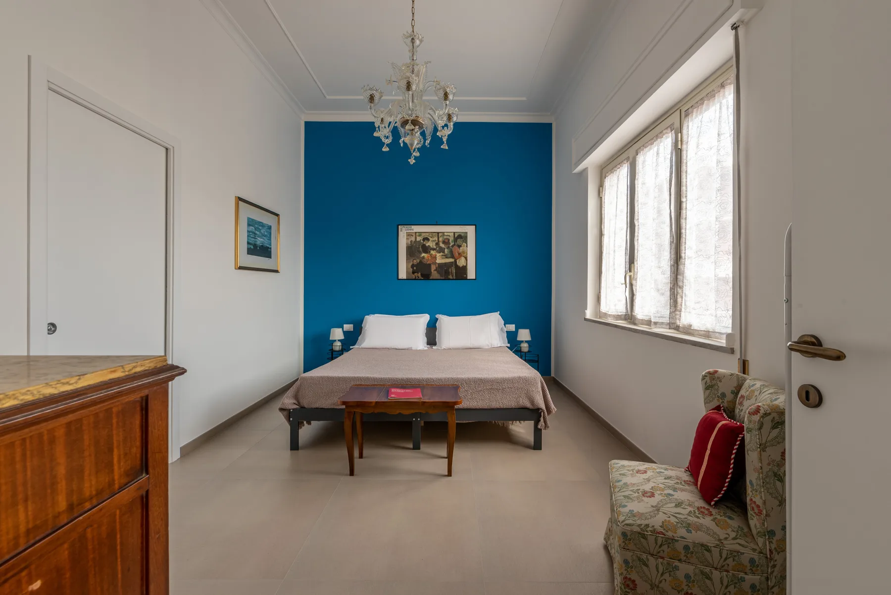 A bedroom with a bed against a blue accent wall and a large window.