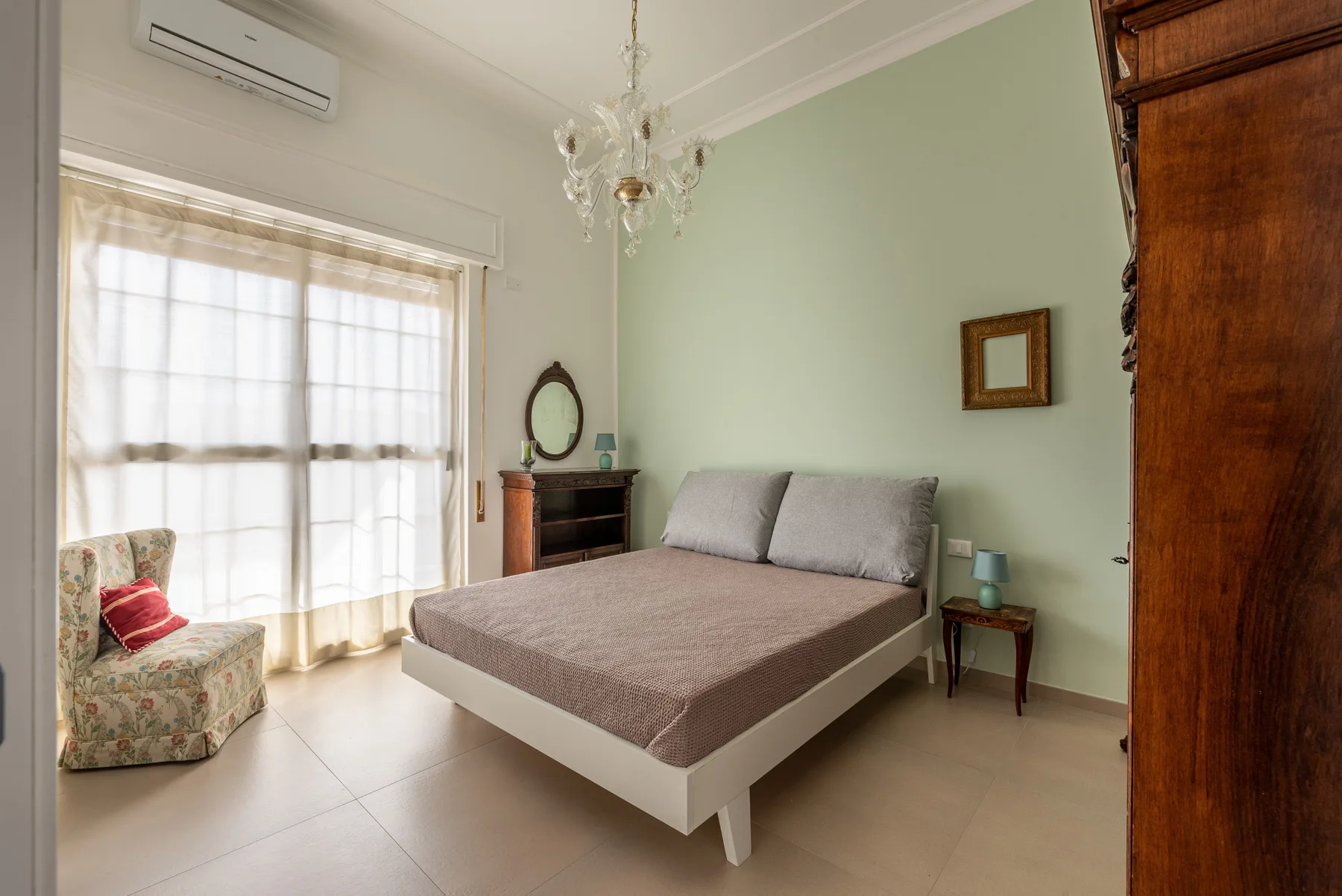A bedroom with a bed beside a large window and an upholstered armchair.
