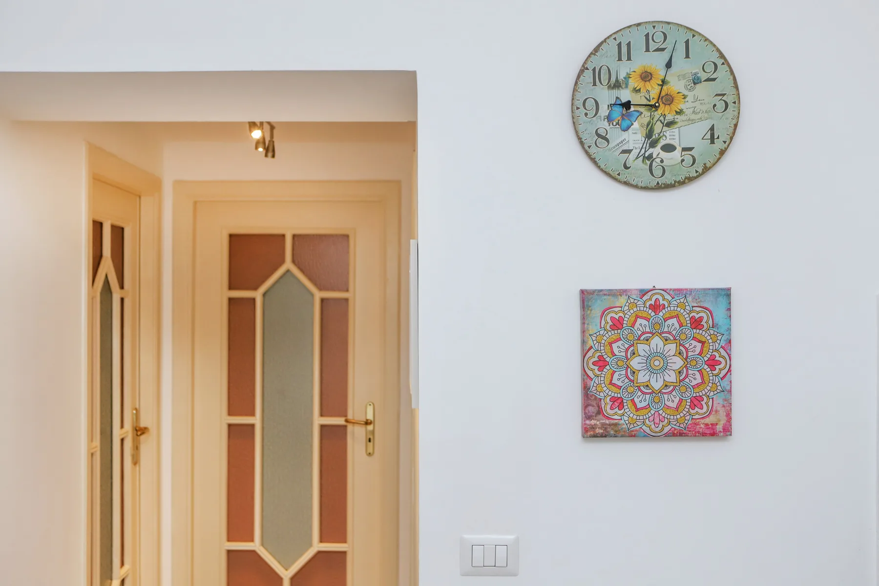 Un pasillo con una puerta decorativa de paneles de vidrio, un reloj y una obra colorida.