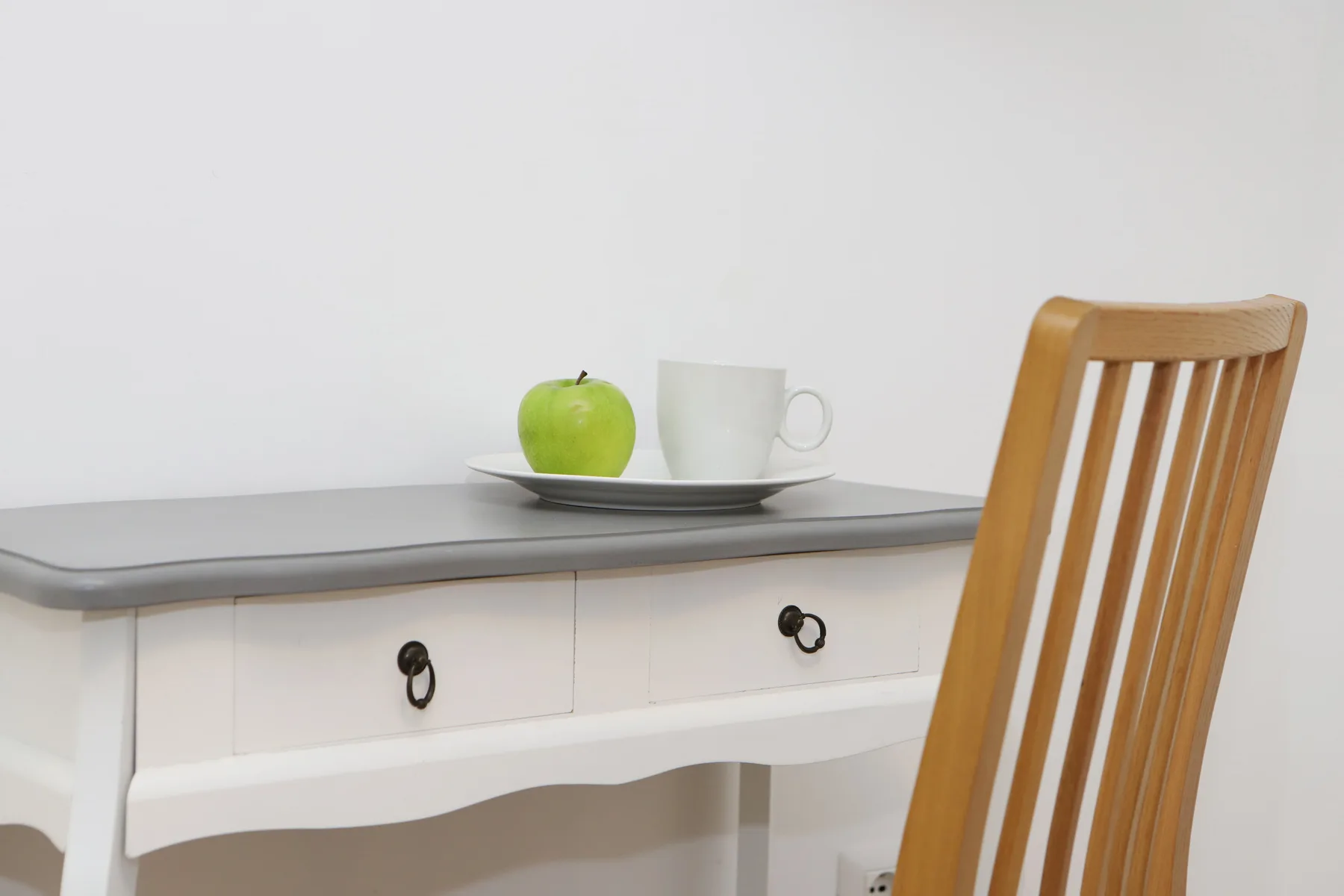 Un escritorio blanco con una silla de madera, una manzana verde y una taza.