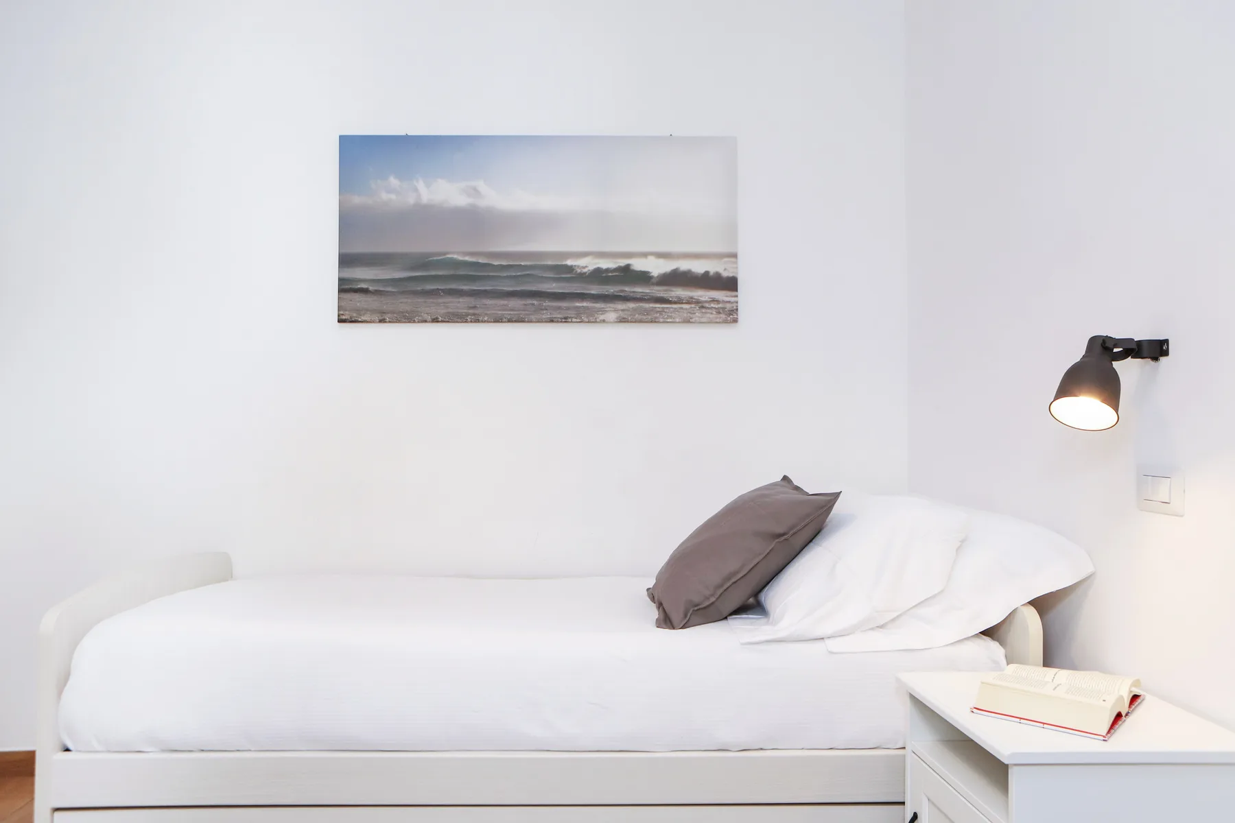 Una cama con ropa de cama blanca está bajo un paisaje marino enmarcado, en una habitación luminosa.