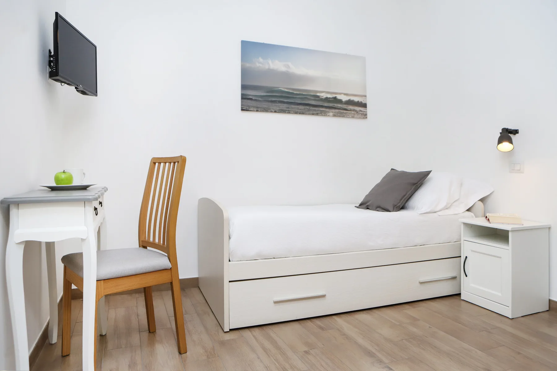 Un dormitorio luminoso con una cama blanca, un escritorio pequeño y un televisor en la pared.
