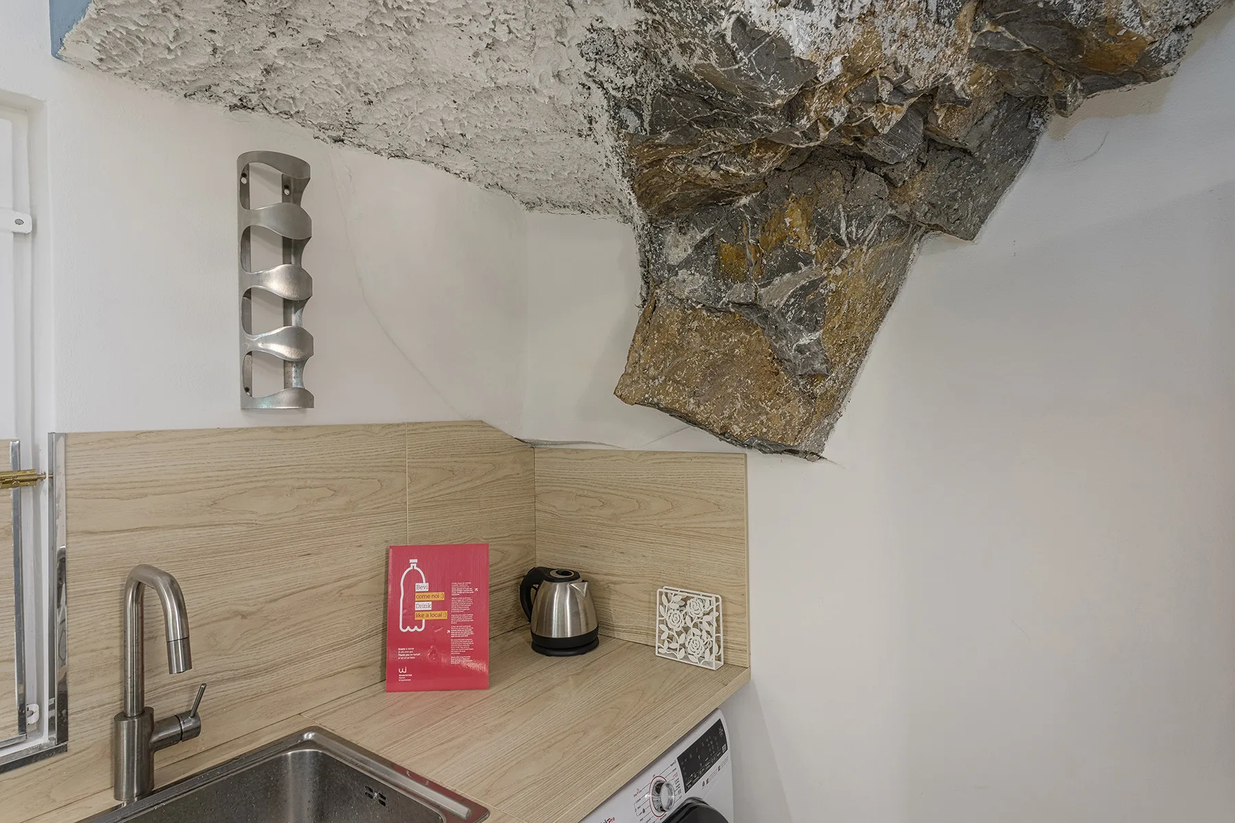 A compact kitchen counter with a sink and kettle sits beneath a rough stone ceiling.
