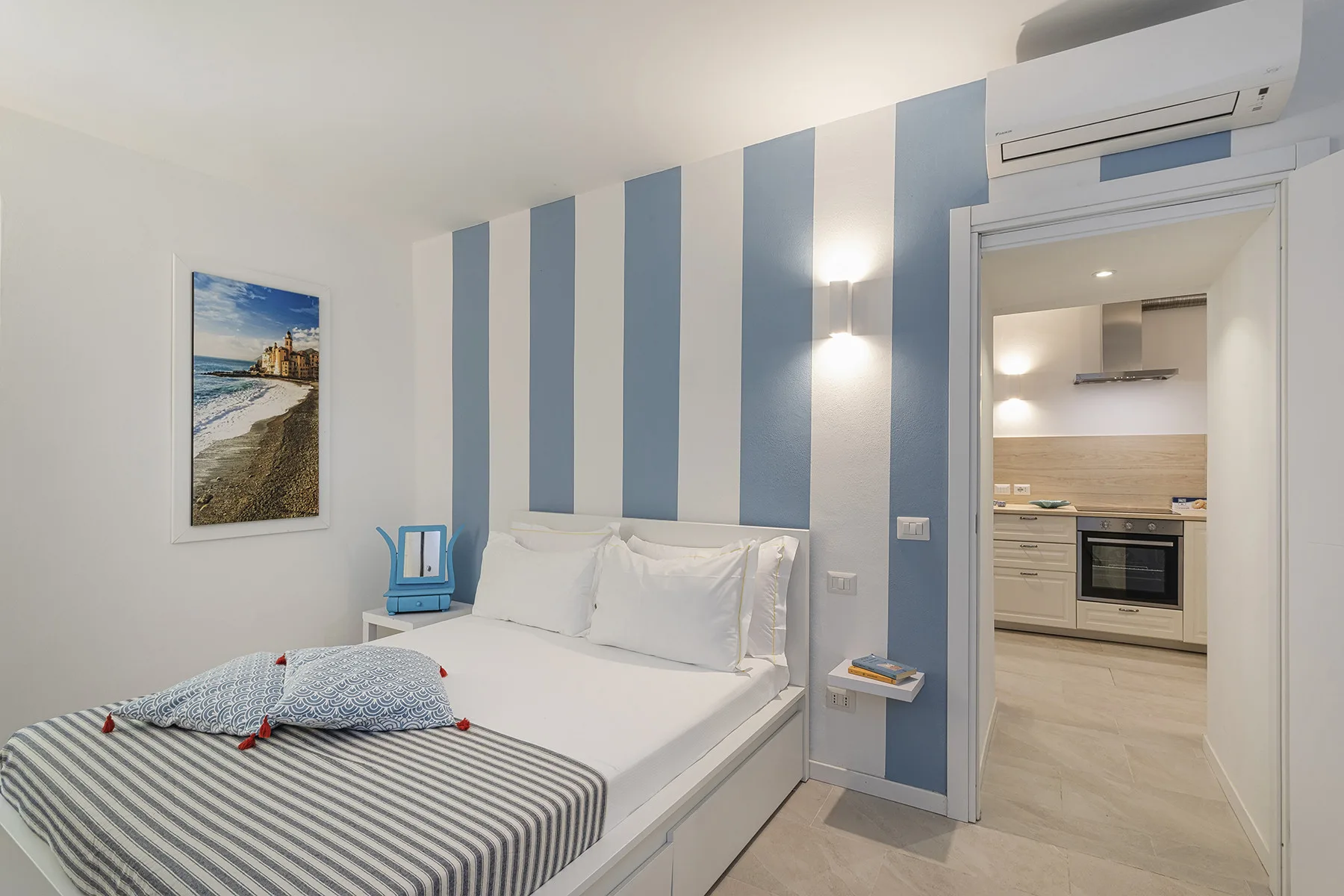 A bedroom with a bed and blue-and-white striped wall beside an open kitchen.