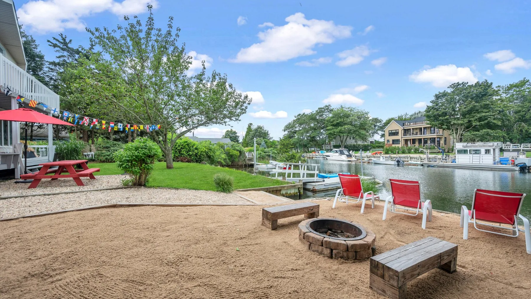 A sandy waterfront seating area has red chairs, a fire pit, and picnic tables.