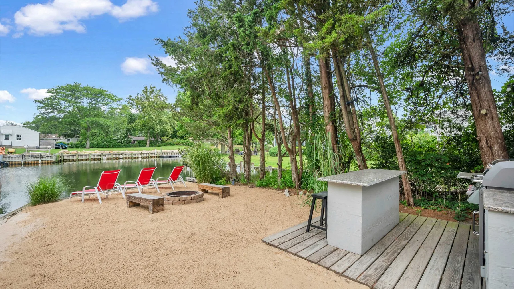 A sandy outdoor seating area beside the water, with red chairs and a small table.
