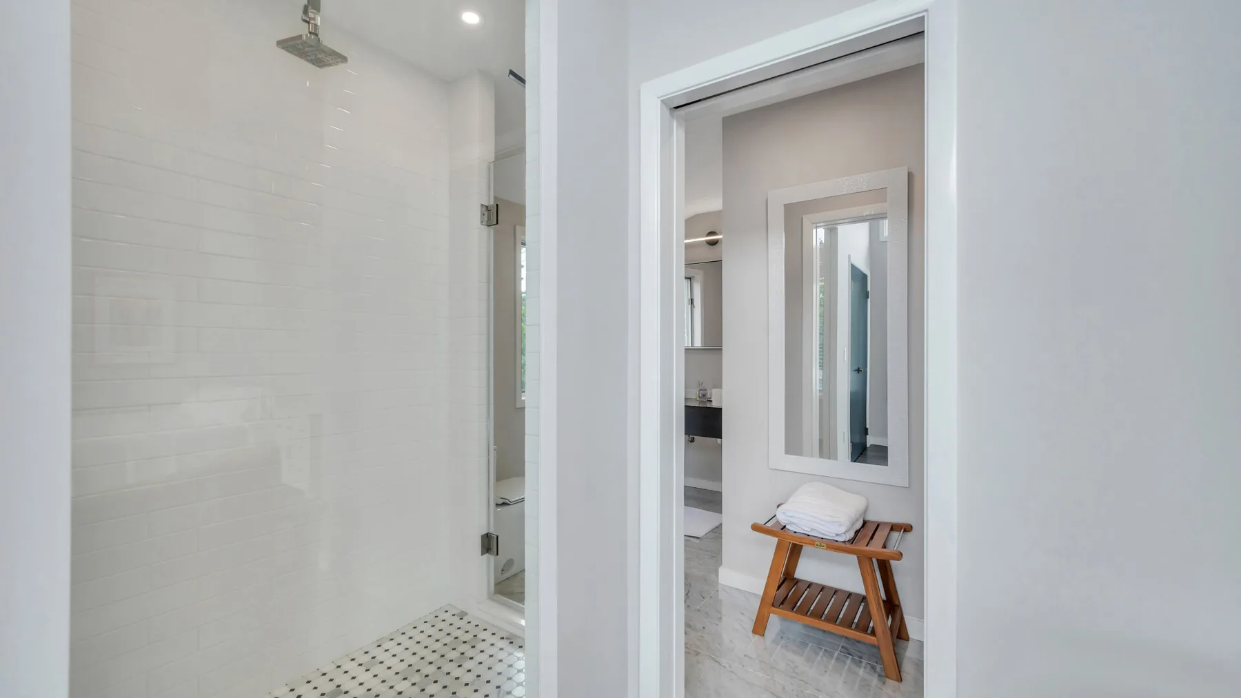 Un baño luminoso con una ducha de vidrio y un taburete de madera junto al lavabo.