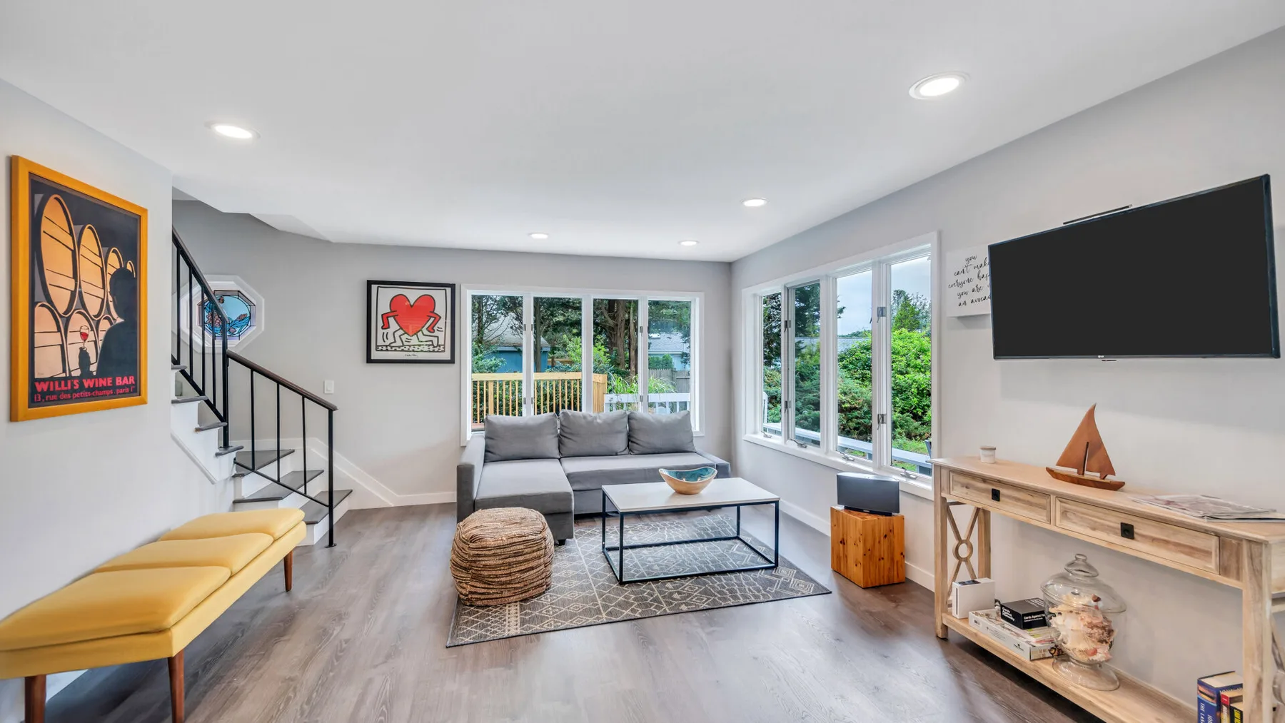 A bright living room with seating, large windows, and a wall-mounted television.