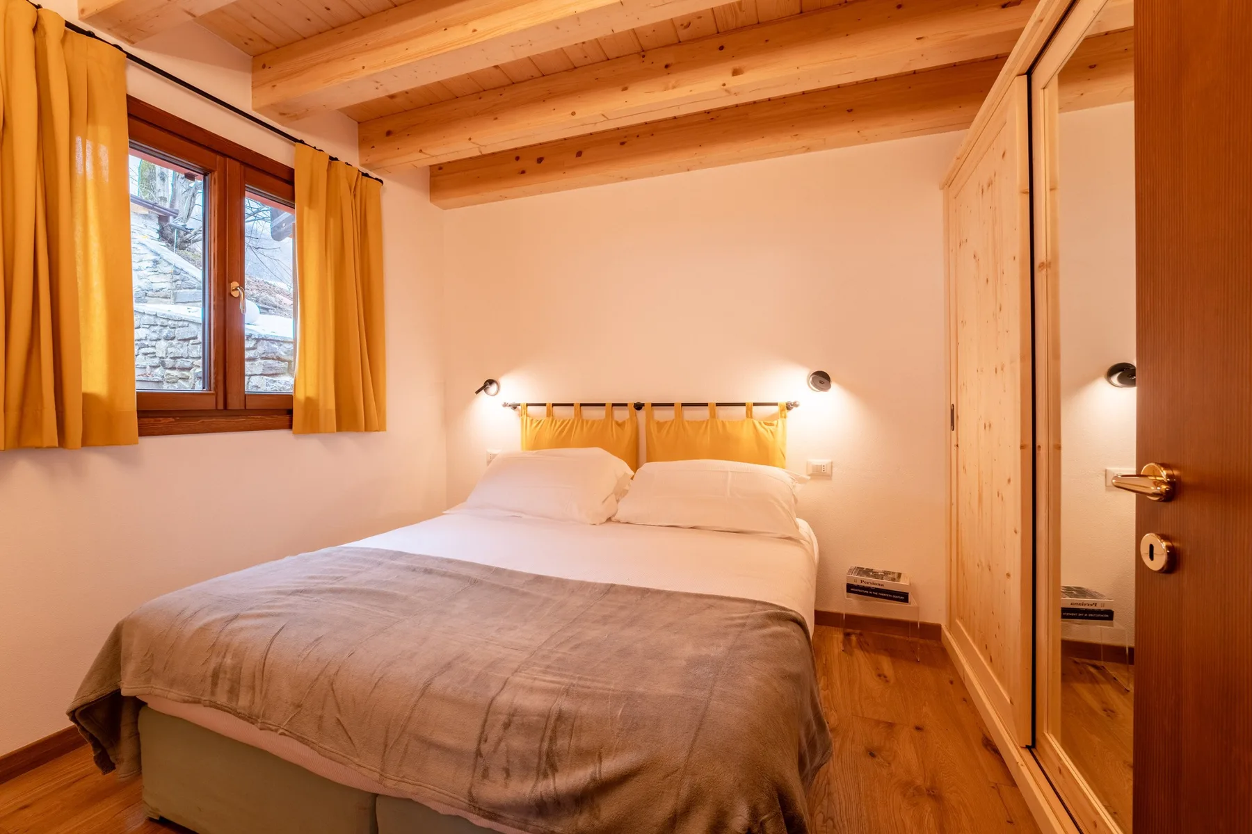A bedroom with a double bed, wooden ceiling beams, and a curtained window.