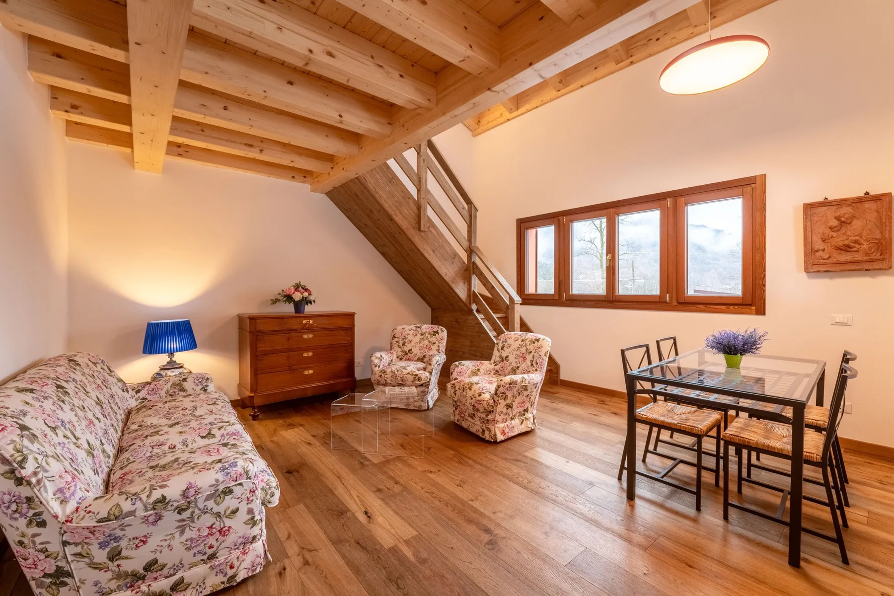 A furnished living room with patterned seating, a wooden staircase, and a large window.