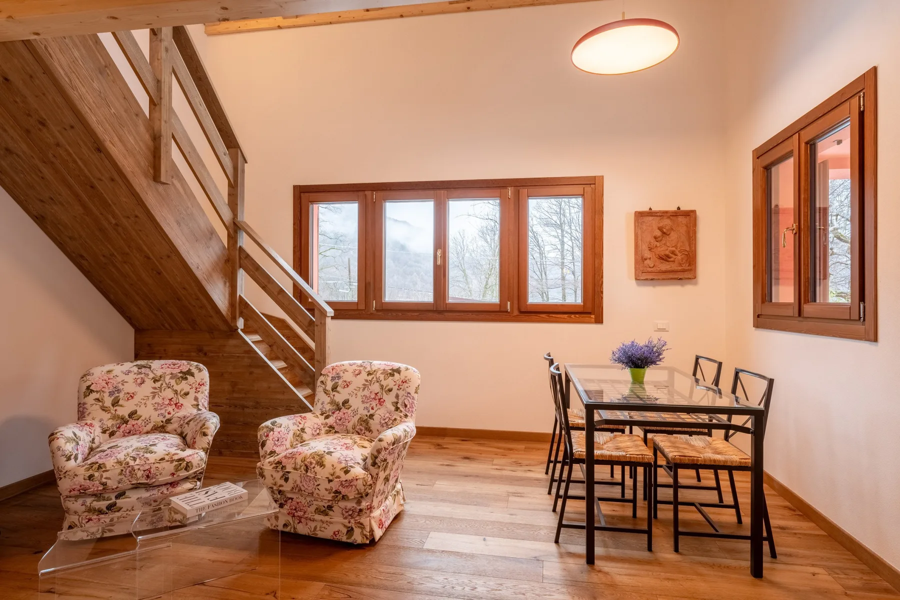 A dining area with a table and chairs beside a wooden staircase and two floral armchairs.