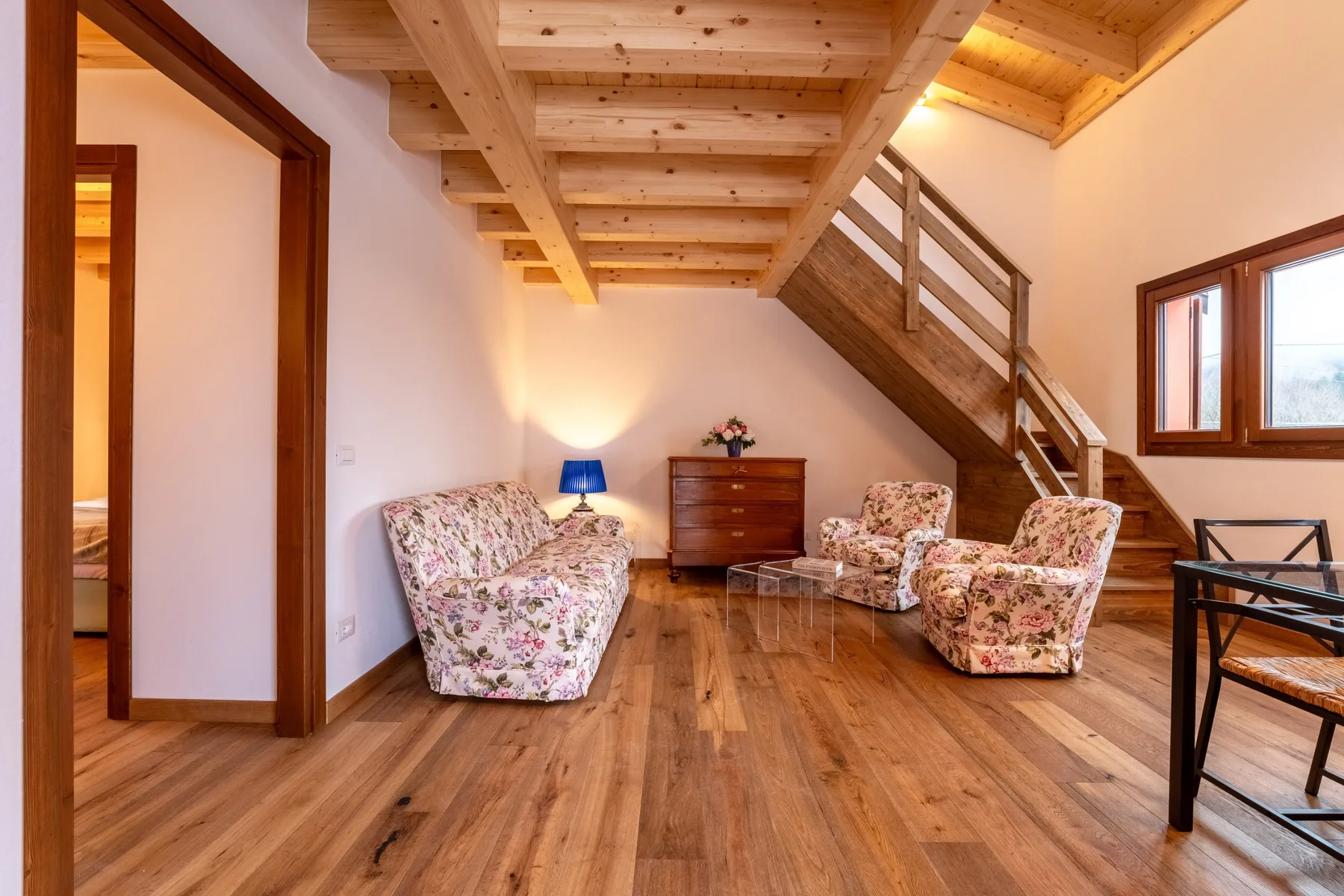A wood-floored sitting area has floral seating beneath a staircase and exposed wooden beams.