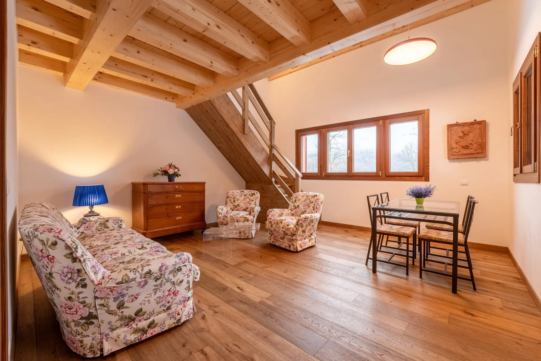 A bright living room with floral seating, a dining table, and a wooden staircase.