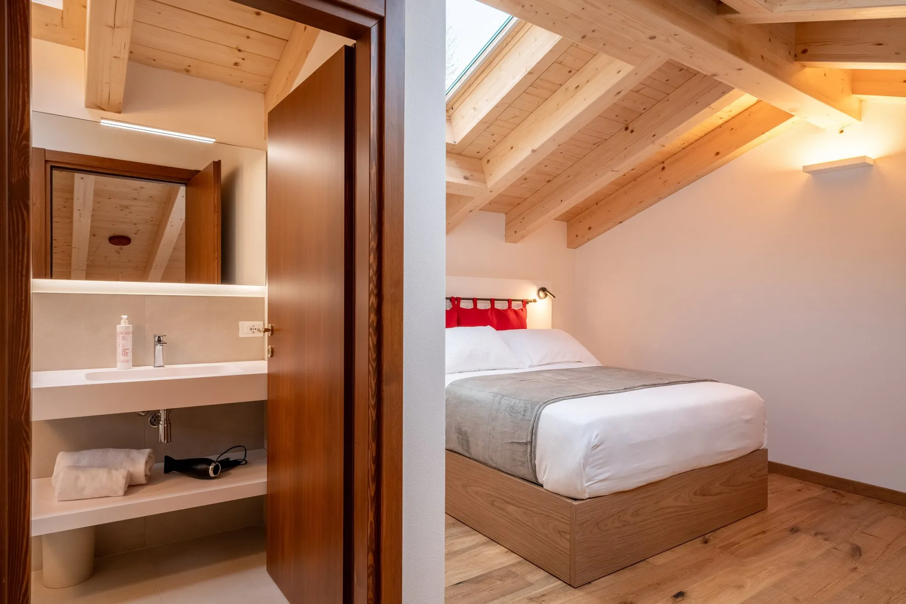 A bedroom with a bed beneath a sloped wooden ceiling, viewed beside an open bathroom vanity.