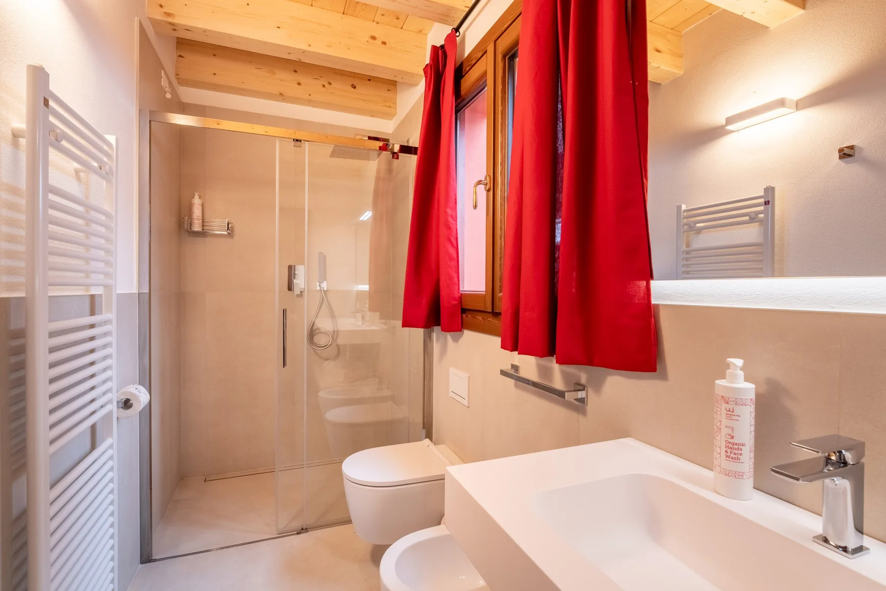 A bathroom with a glass shower, sink, and red curtains by the window.