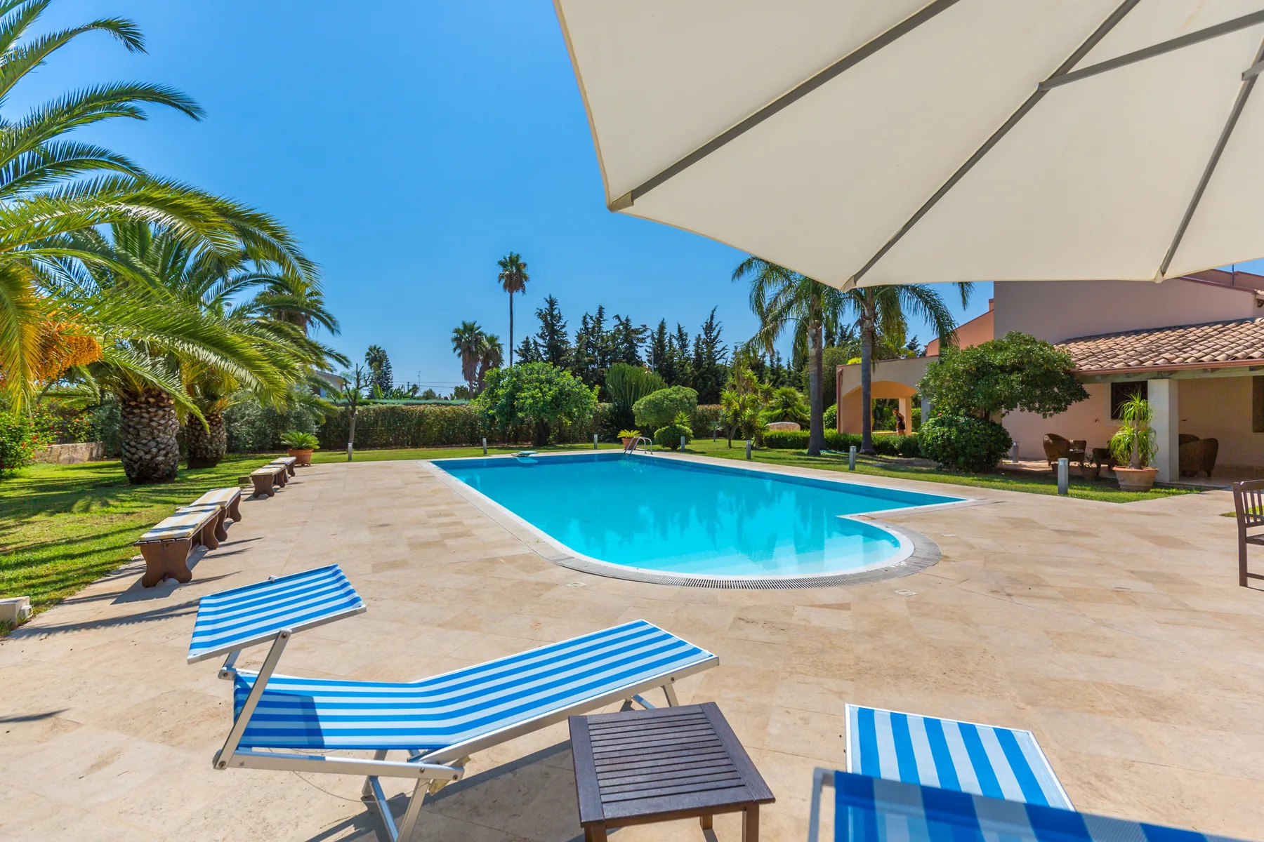 An outdoor swimming pool is surrounded by a paved deck, lounge seating, and greenery.