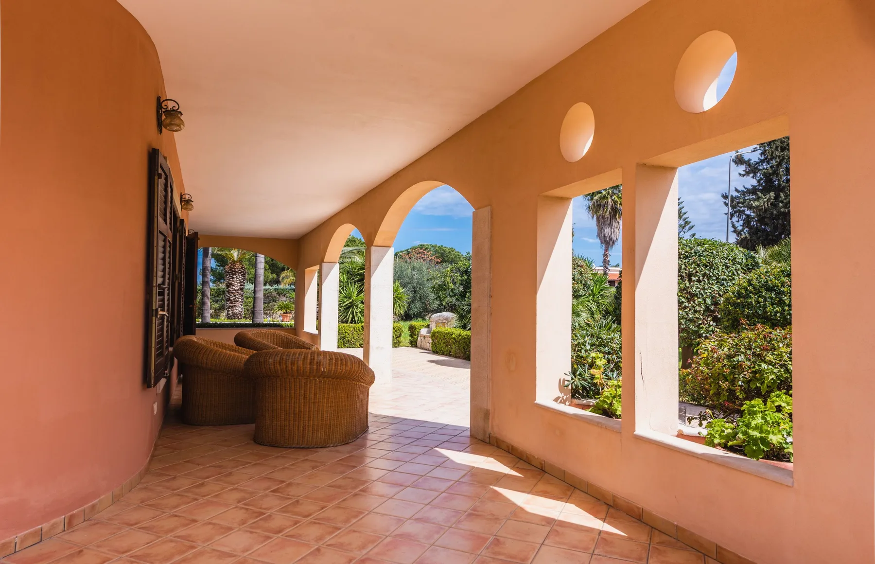 Un patio cubierto con aberturas arqueadas, suelo de baldosas y asientos de mimbre da a un jardín.