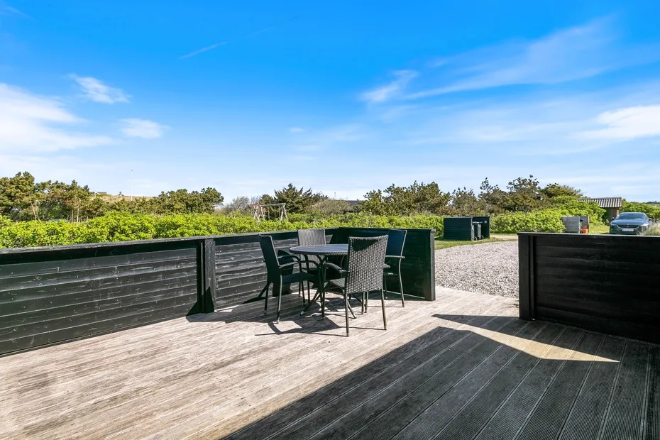 Una terraza exterior con mesa y asientos da a una zona verde bajo un cielo azul.