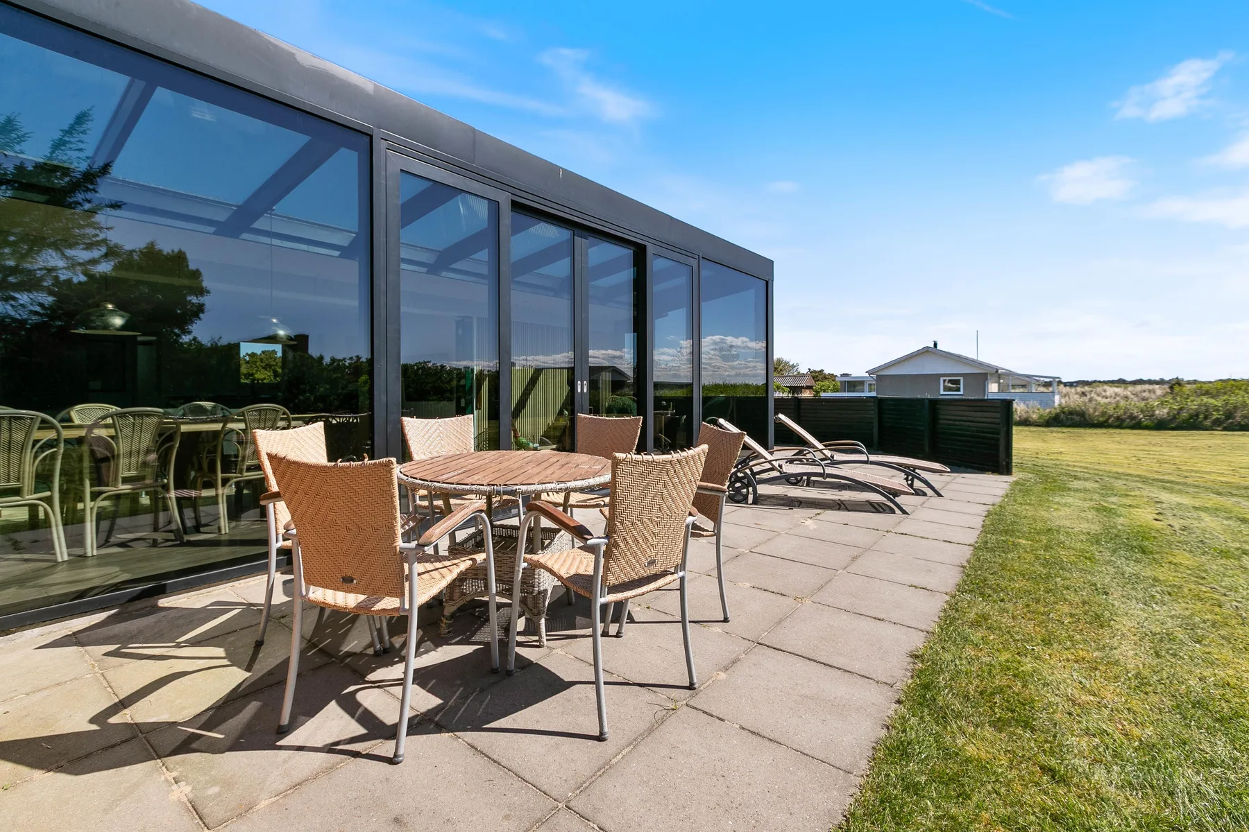 Un patio exterior con mesa y asientos junto a grandes ventanas de vidrio y una zona de césped.