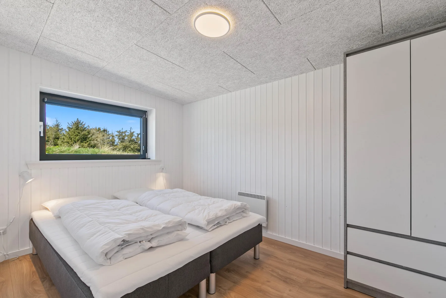 Un dormitorio luminoso con una cama doble junto a una ventana con vistas a los árboles.