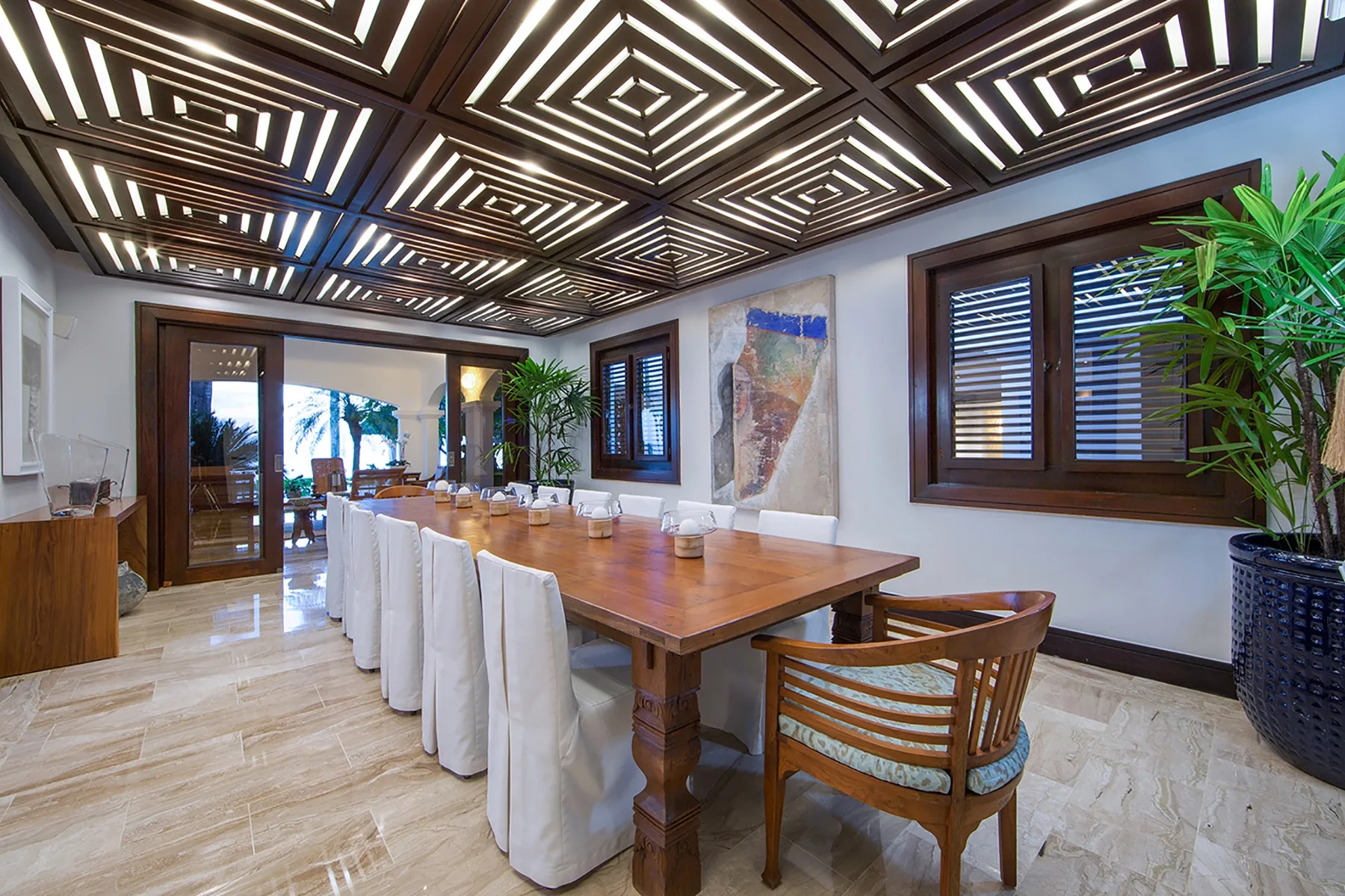 A dining room with a long wooden table and chairs beneath a patterned ceiling.