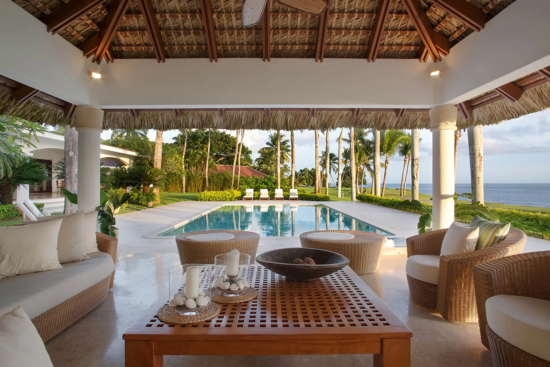 An open-sided covered patio overlooks a pool, tropical garden, and ocean.