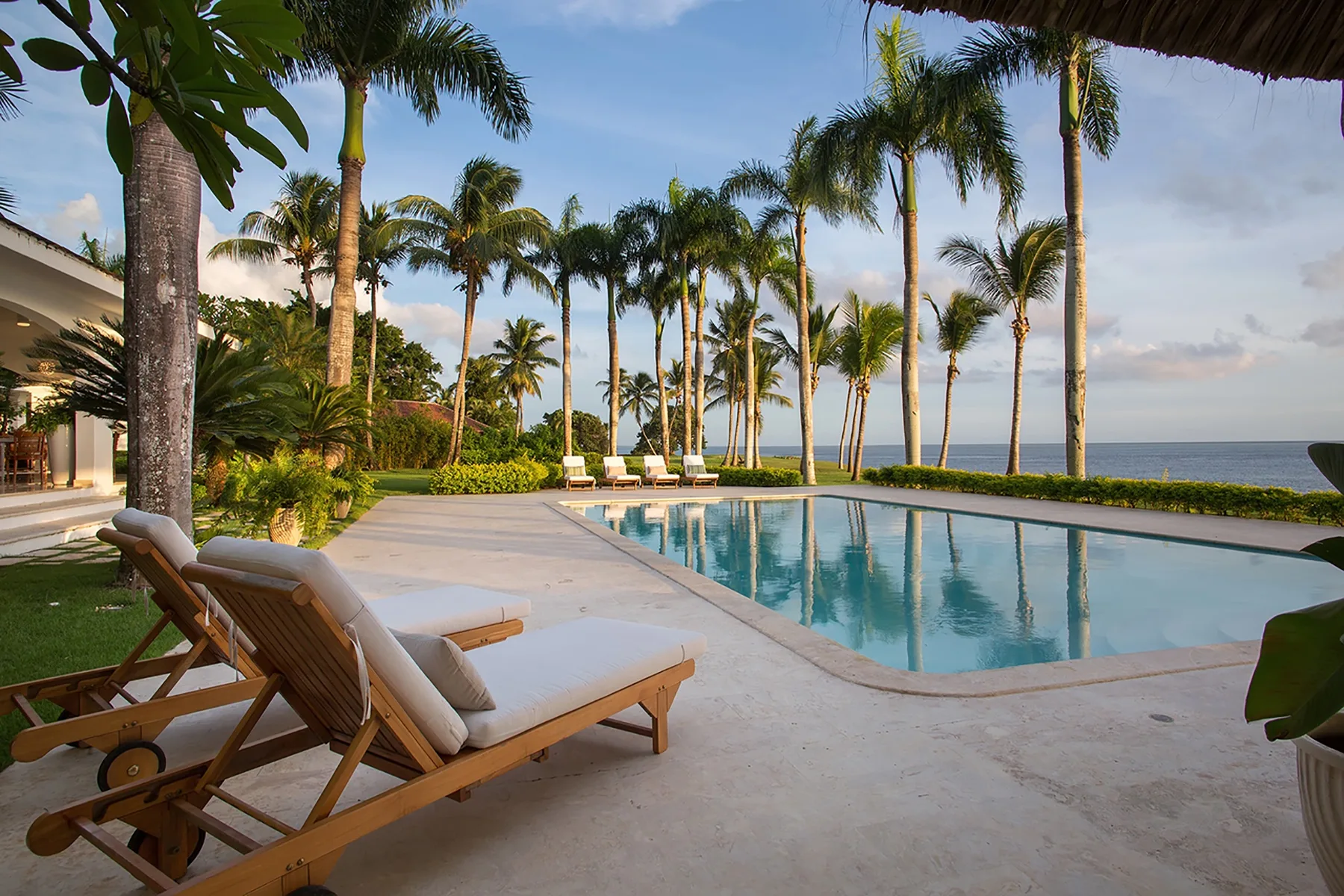 A swimming pool beside lounge seating, palm trees, and the ocean.