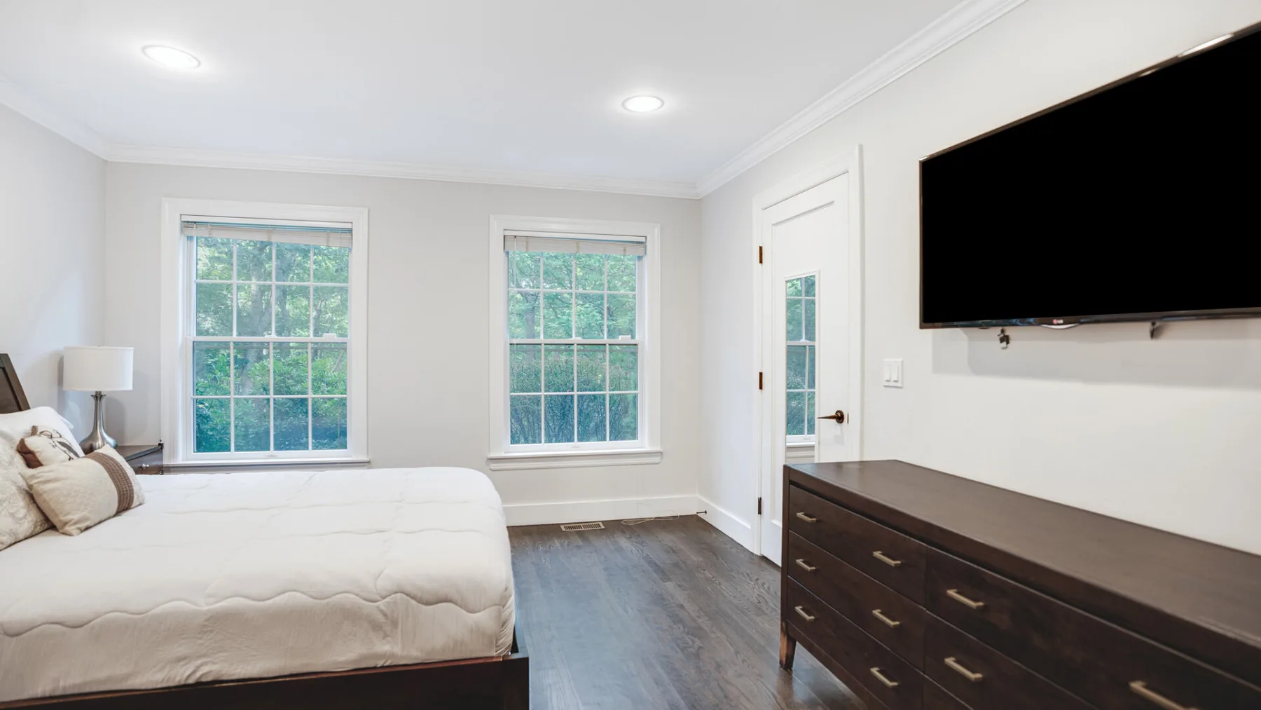 Un dormitorio luminoso con una cama, dos ventanas y una cómoda bajo un televisor montado en la pared.