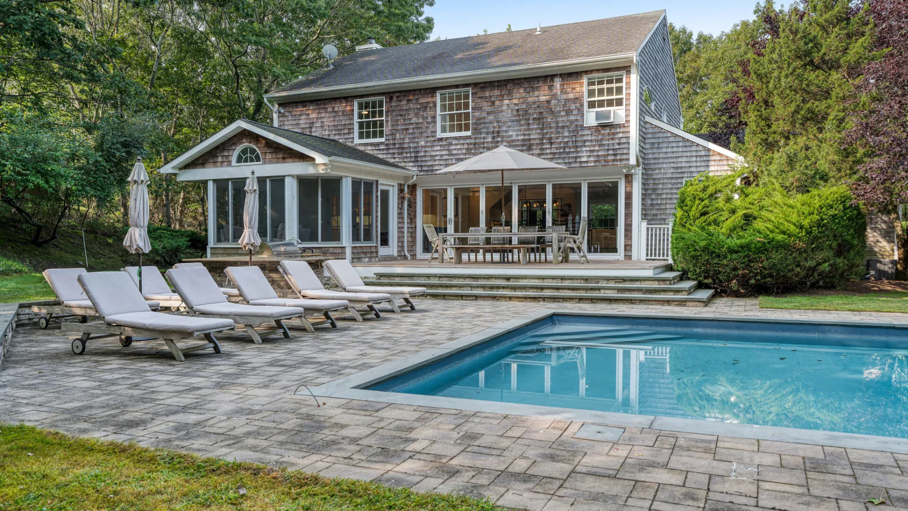 Una piscina y un patio pavimentado se encuentran junto a una casa rodeada de árboles.
