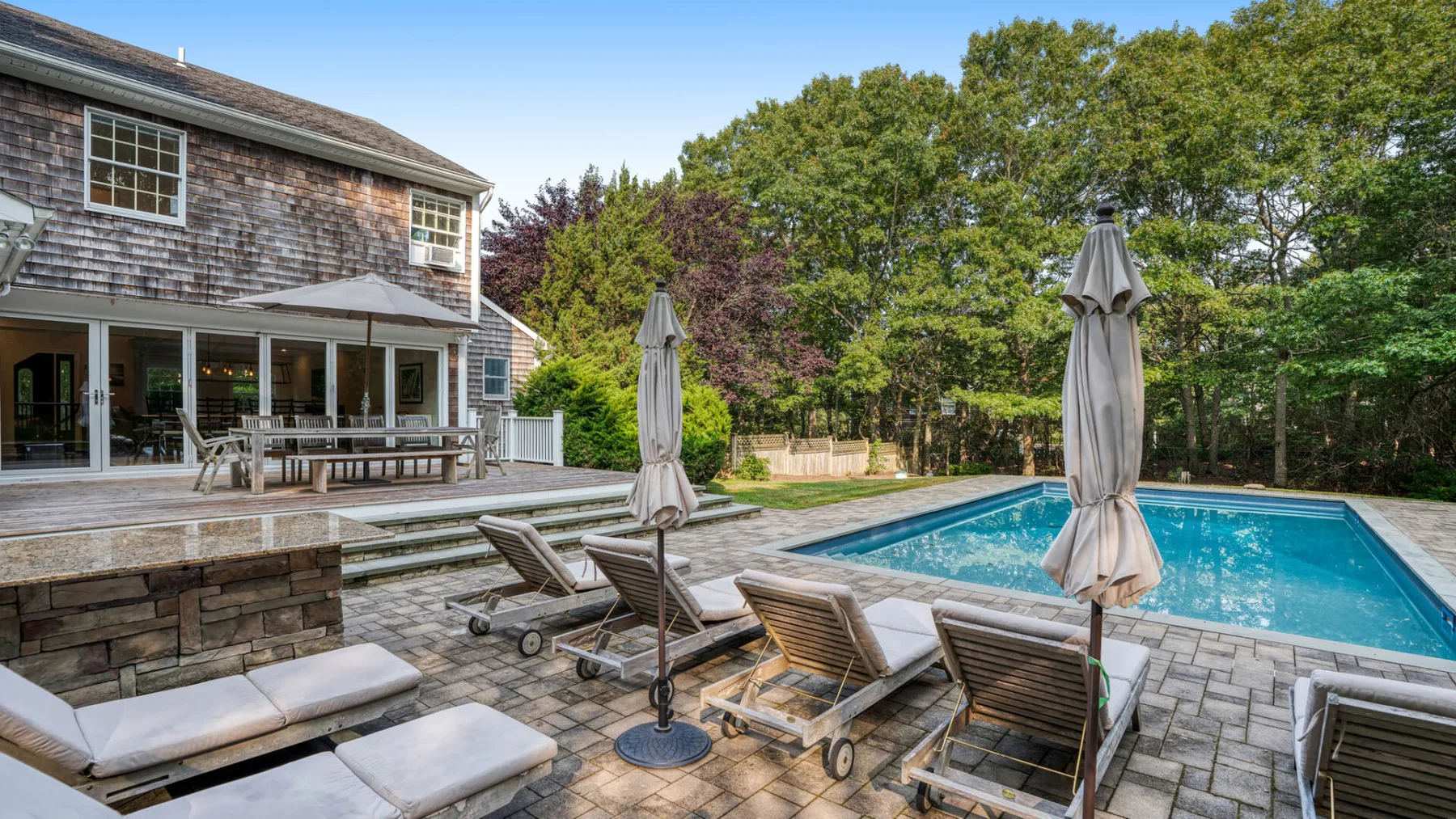 Una piscina exterior y un patio pavimentado junto a una casa, con tumbonas y sombrillas cerradas.