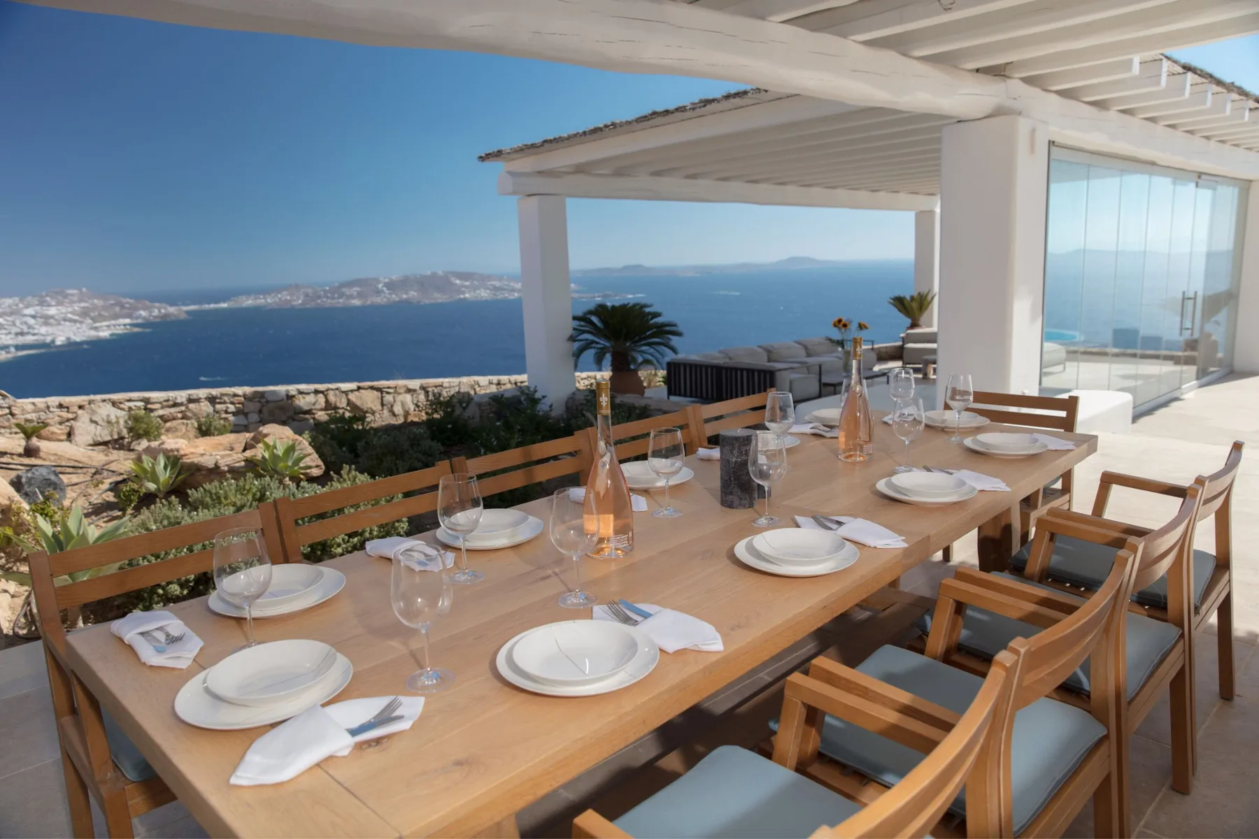 An outdoor dining table is set beneath a white pergola overlooking the sea.