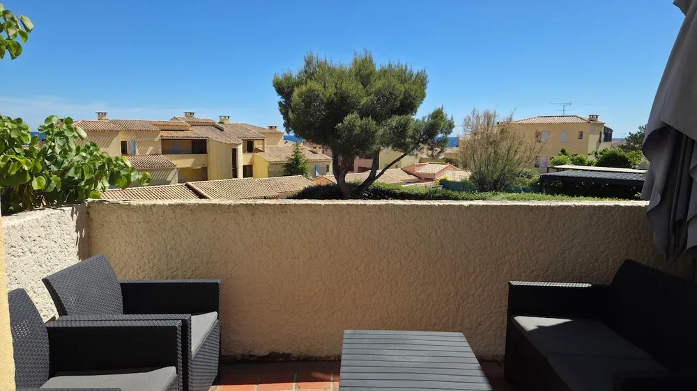 A sunny terrace with dark seating overlooks nearby houses and trees.