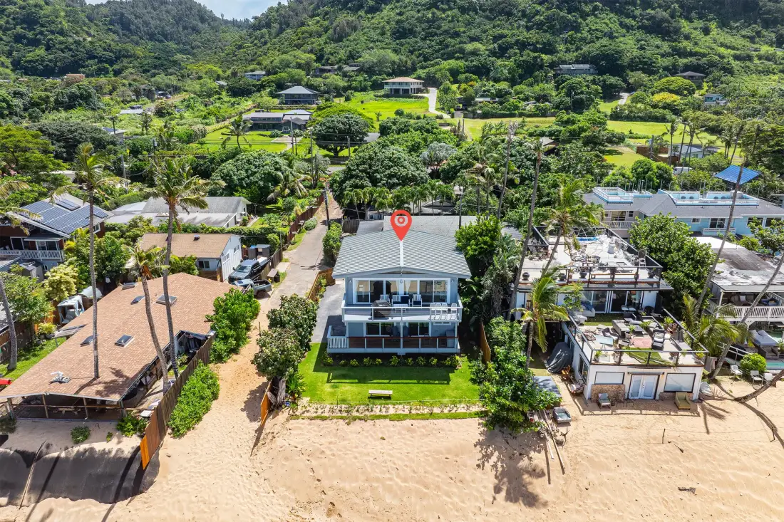 Vista aérea de una casa frente a la playa con un pueblo en una ladera verde detrás.