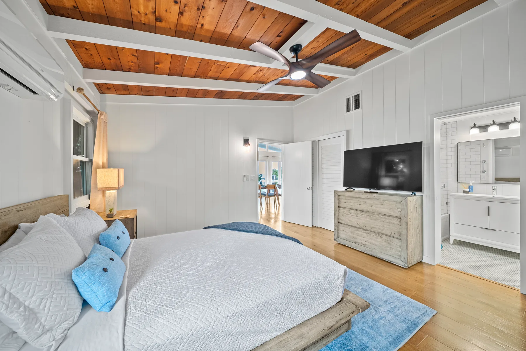Un dormitorio luminoso con una cama, ventilador de techo y televisor sobre una cómoda.