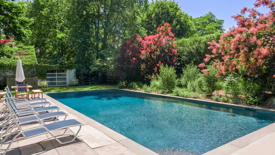 An outdoor swimming pool is bordered by lounge seating and flowering trees.