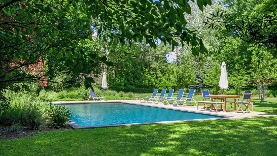An outdoor swimming pool is surrounded by lawn, trees, and lounge seating.