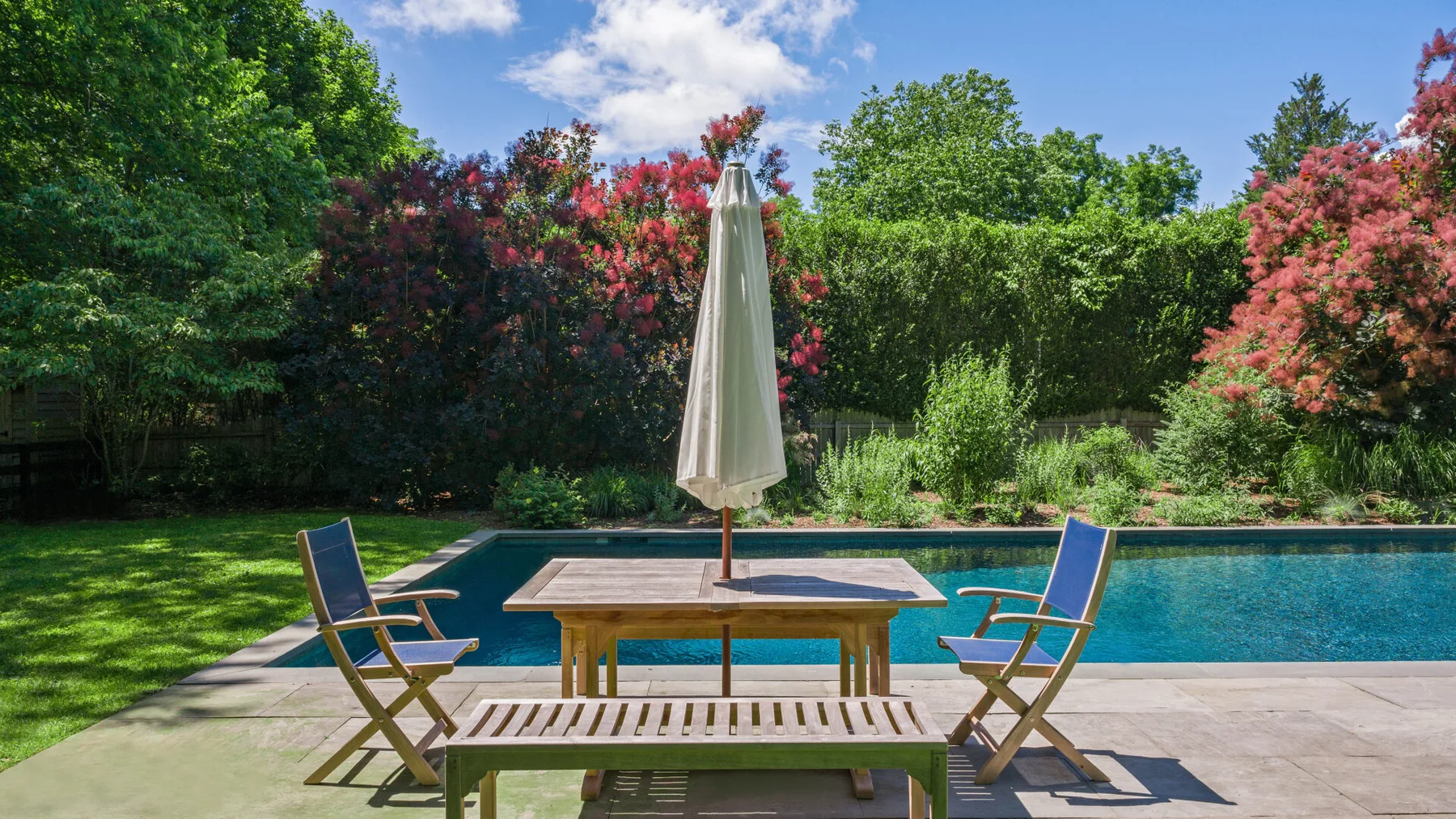 A poolside patio has a table, seating, and a closed umbrella.
