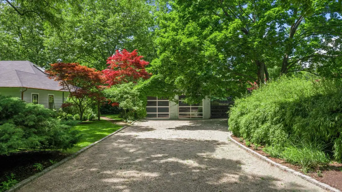 A gravel driveway leads to a gate, surrounded by trees and shrubs.