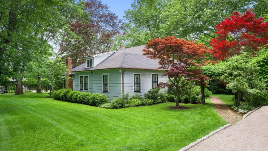 A small house sits beside a broad green lawn, surrounded by leafy trees.