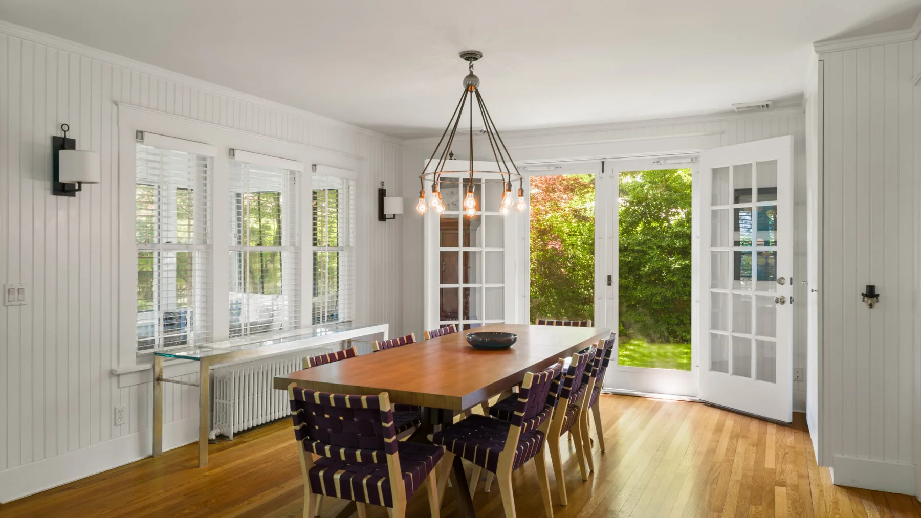A bright dining room with a long wooden table and open glass doors.