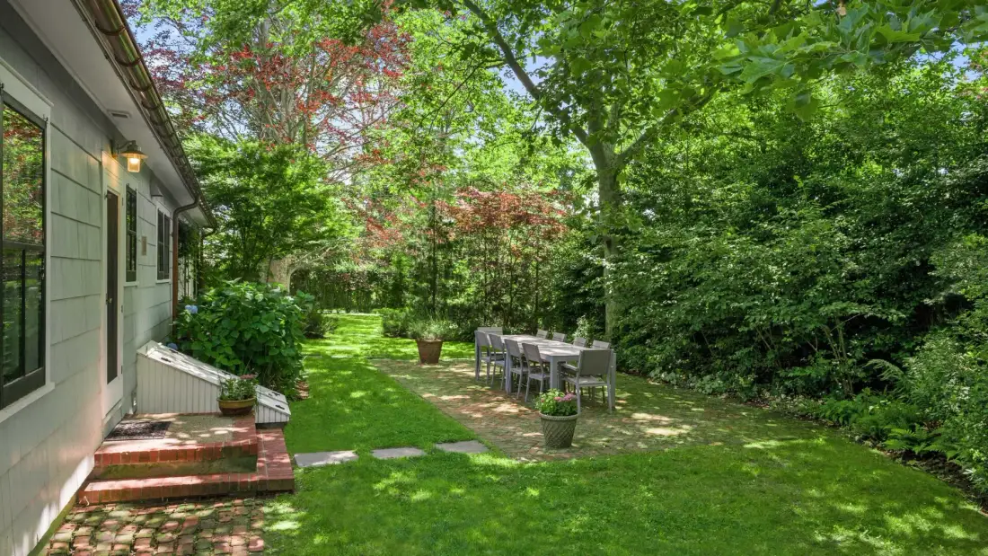 A garden patio beside a house, with outdoor seating under leafy trees.