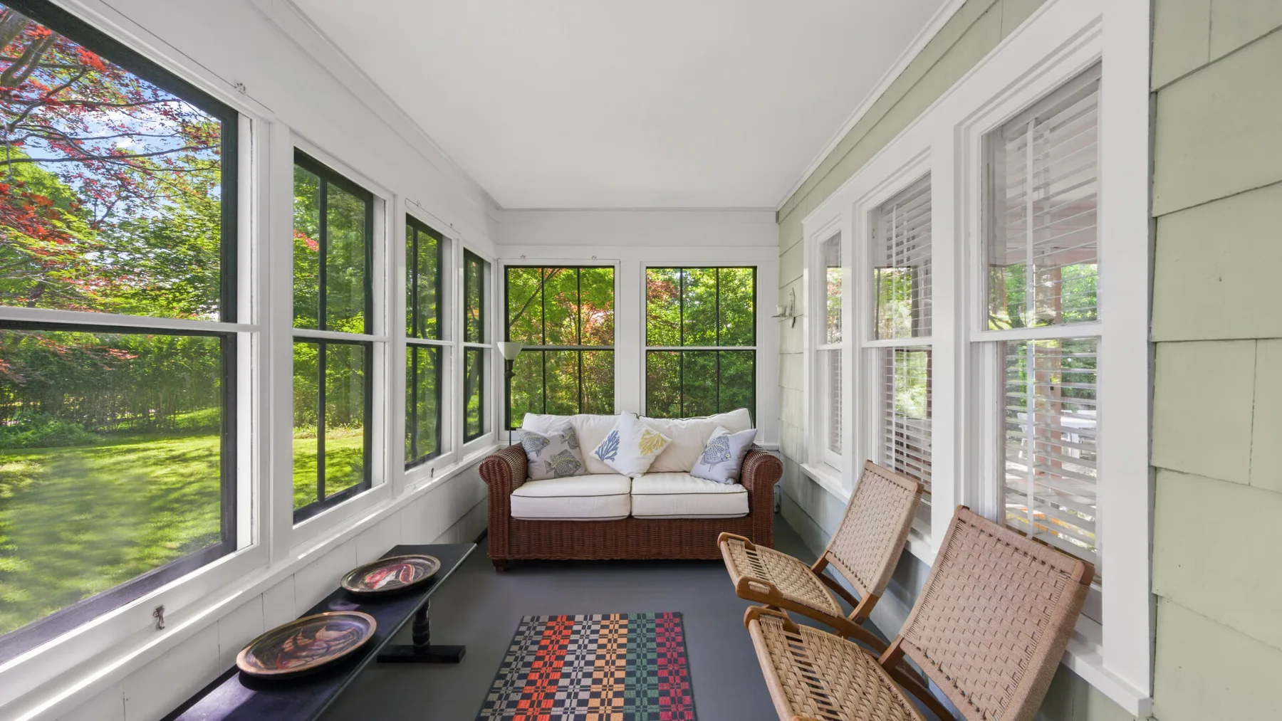 A bright enclosed sunroom with seating and large windows overlooking greenery.