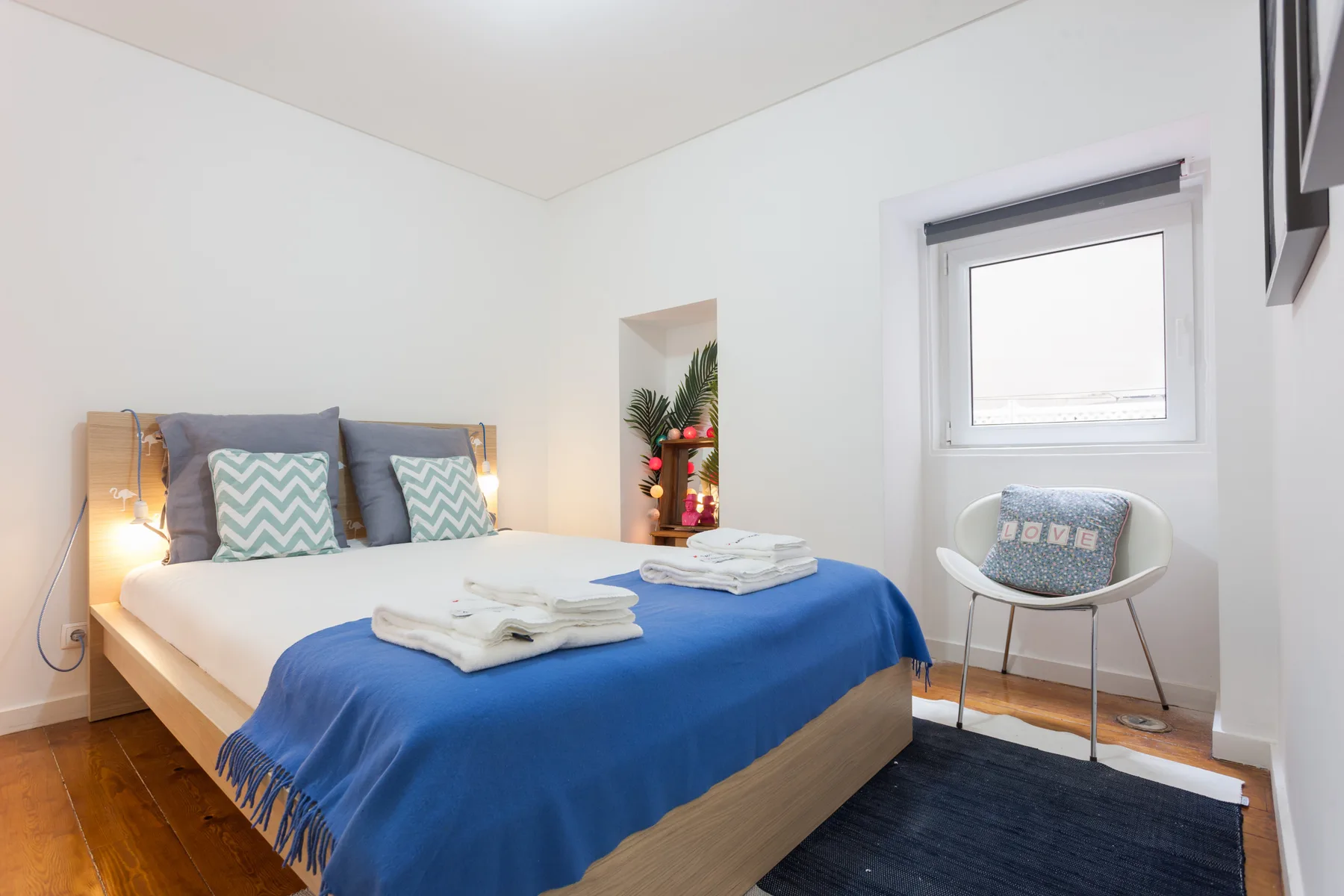Dormitorio con una cama cubierta con ropa de cama azul y una silla junto a la ventana.