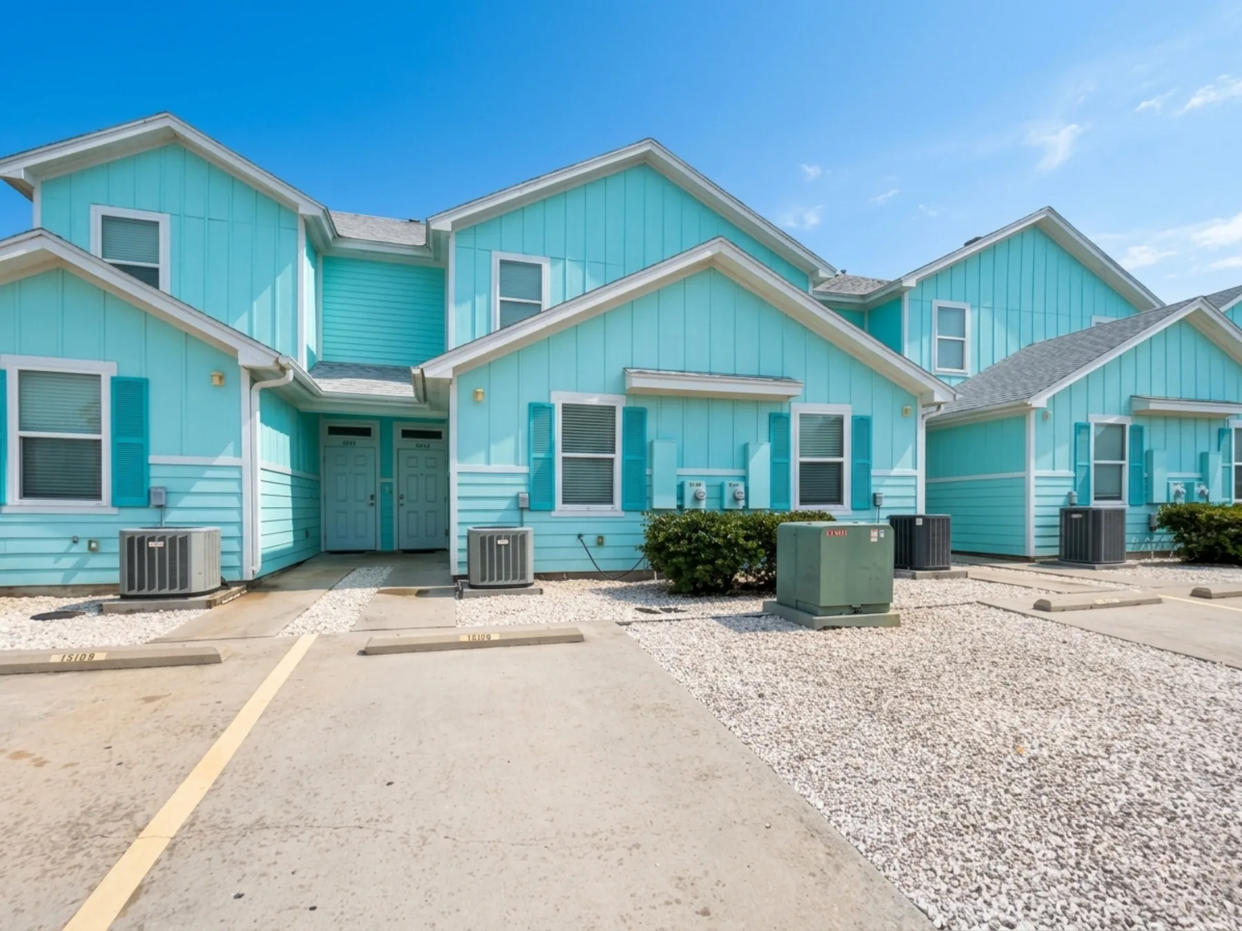 A row of turquoise residential buildings faces a paved parking area.