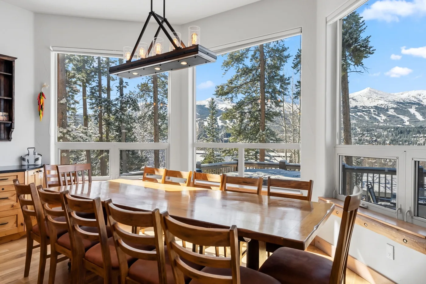 Un comedor con una larga mesa de madera junto a grandes ventanas con vistas a las montañas.