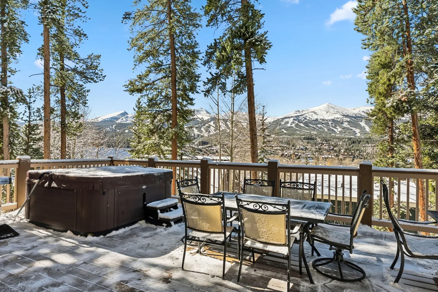 Una terraza exterior con una bañera de hidromasaje y una mesa de comedor da a un paisaje montañoso nevado.