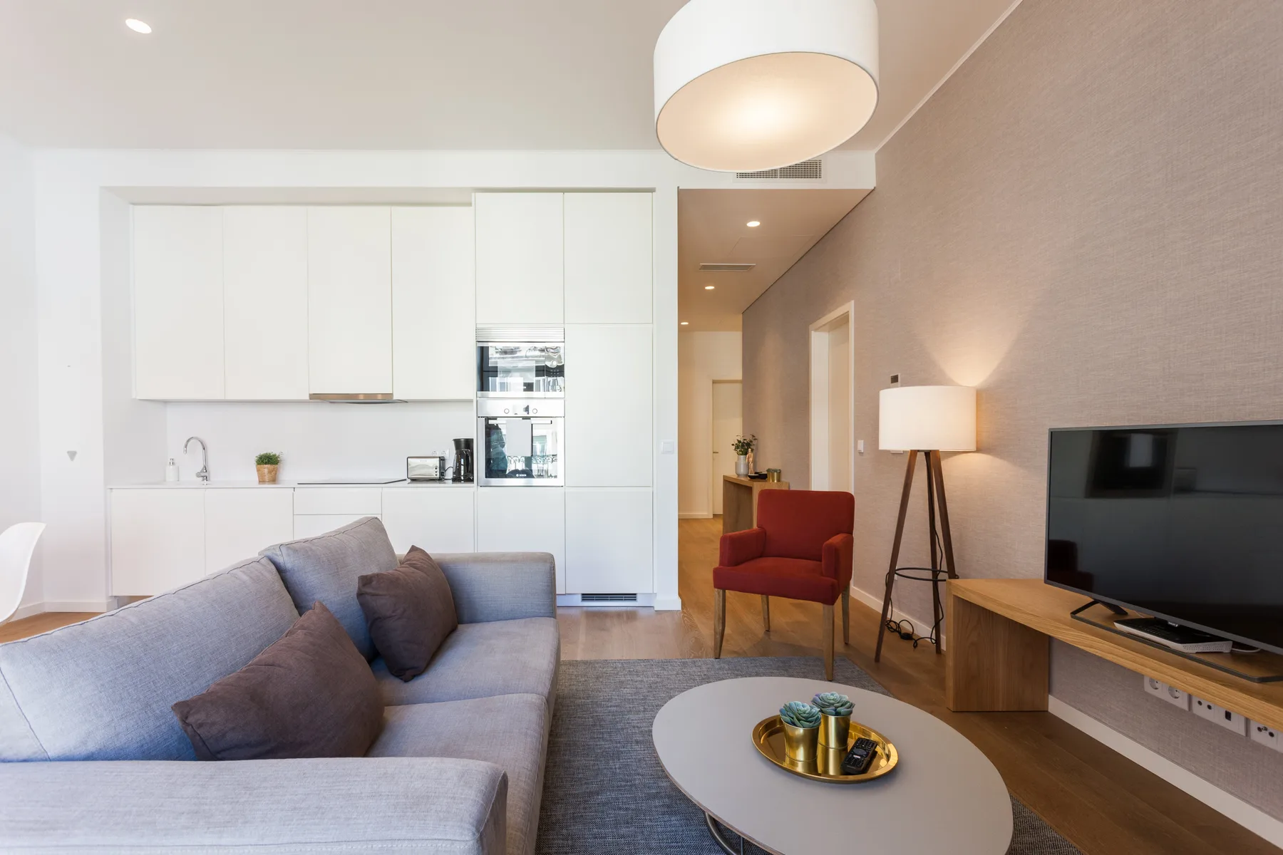 Una sala luminosa con sofá gris, mesa de centro y televisor junto a la cocina.